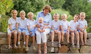 Gammelmormor eller gammelfarmor sitter med ett leende och håller ett litet barn. Runtomkring sitter åtta barn i vita tröjor och blå shorts, alla glada och förväntansfulla.
