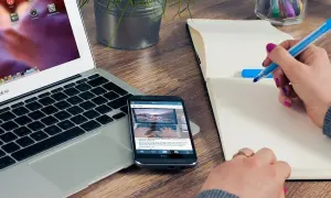 Person skriver i anteckningsbok vid laptop och mobil. Ett bra exempel på en problemformulering kan vara att hitta effektiva metoder för distansarbete.