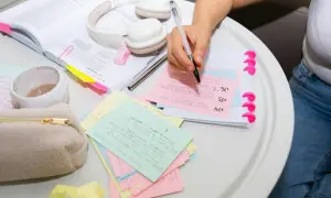 Person skriver på studieflikskort, omgiven av böcker och hörlurar. Kort med texten "dem andra" är synliga.