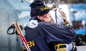 Två skidåkare i svenska färger kramas. Mannen har mössa med texten "SWE" och kvinnan har pannband. De står fram emot varandra i en varm omfamning.