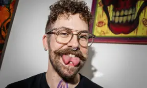 Man med glasögon, mustasch och septumring visar upp sin kluvna tunga. Han verkar tala med kluven tunga, vilket ger ett unikt uttryck.