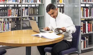 Man sitter vid ett runt bord i ett bibliotek, med en laptop och böcker. Han läser i en bok och funderar på hur citerar man.