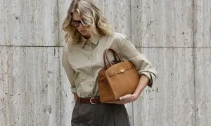 En kvinna med blont hår och glasögon håller en brun handväska. Hon bär en beige skjorta och mörkbruna byxor. Om hon ska på möte eller bara ut på stan är oklart.