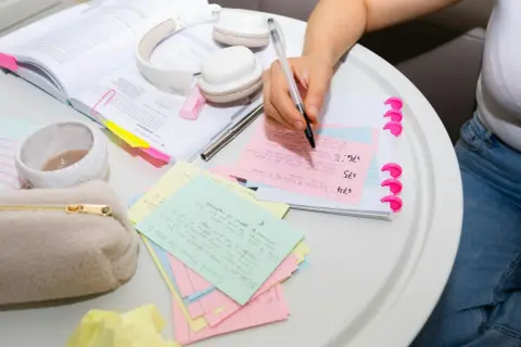 Person skriver på studieflikskort, omgiven av böcker och hörlurar. Kort med texten "dem andra" är synliga.