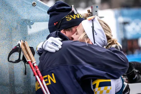 Två skidåkare i svenska färger kramas. Mannen har mössa med texten "SWE" och kvinnan har pannband. De står fram emot varandra i en varm omfamning.