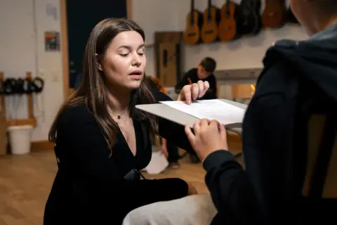 En lärare förklarar hur man skriver en rapport för en elev. I bakgrunden hänger gitarrer.