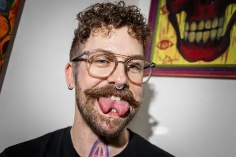 Man med glasögon, mustasch och septumring visar upp sin kluvna tunga. Han verkar tala med kluven tunga, vilket ger ett unikt uttryck.