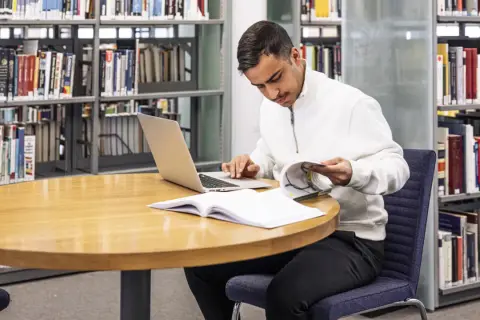 Man sitter vid ett runt bord i ett bibliotek, med en laptop och böcker. Han läser i en bok och funderar på hur citerar man.