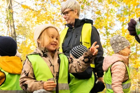Barn i reflexvästar och mössor i höstskogen. En vuxen deltar i leken med en flaska.