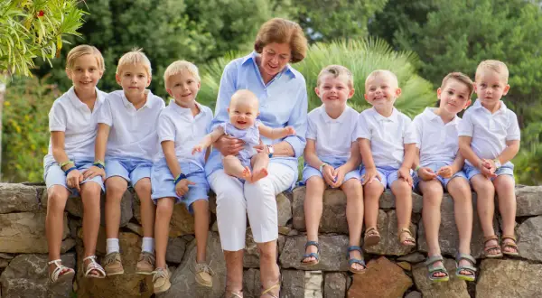 Gammelmormor eller gammelfarmor sitter med ett leende och håller ett litet barn. Runtomkring sitter åtta barn i vita tröjor och blå shorts, alla glada och förväntansfulla.