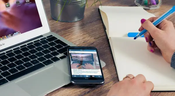 Person skriver i anteckningsbok vid laptop och mobil. Ett bra exempel på en problemformulering kan vara att hitta effektiva metoder för distansarbete.