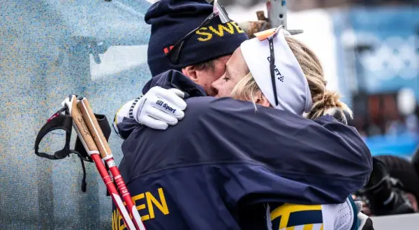 Två skidåkare i svenska färger kramas. Mannen har mössa med texten "SWE" och kvinnan har pannband. De står fram emot varandra i en varm omfamning.