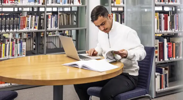 Man sitter vid ett runt bord i ett bibliotek, med en laptop och böcker. Han läser i en bok och funderar på hur citerar man.