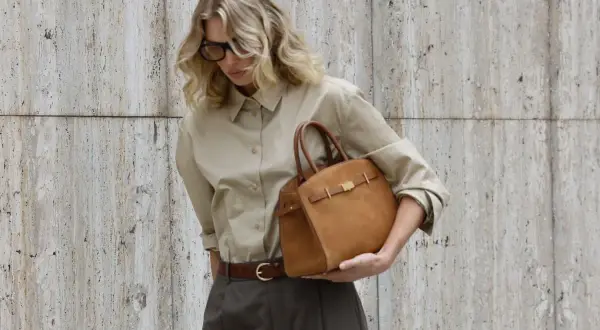 En kvinna med blont hår och glasögon håller en brun handväska. Hon bär en beige skjorta och mörkbruna byxor. Om hon ska på möte eller bara ut på stan är oklart.