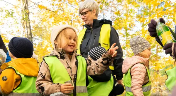 Barn i reflexvästar och mössor i höstskogen. En vuxen deltar i leken med en flaska.