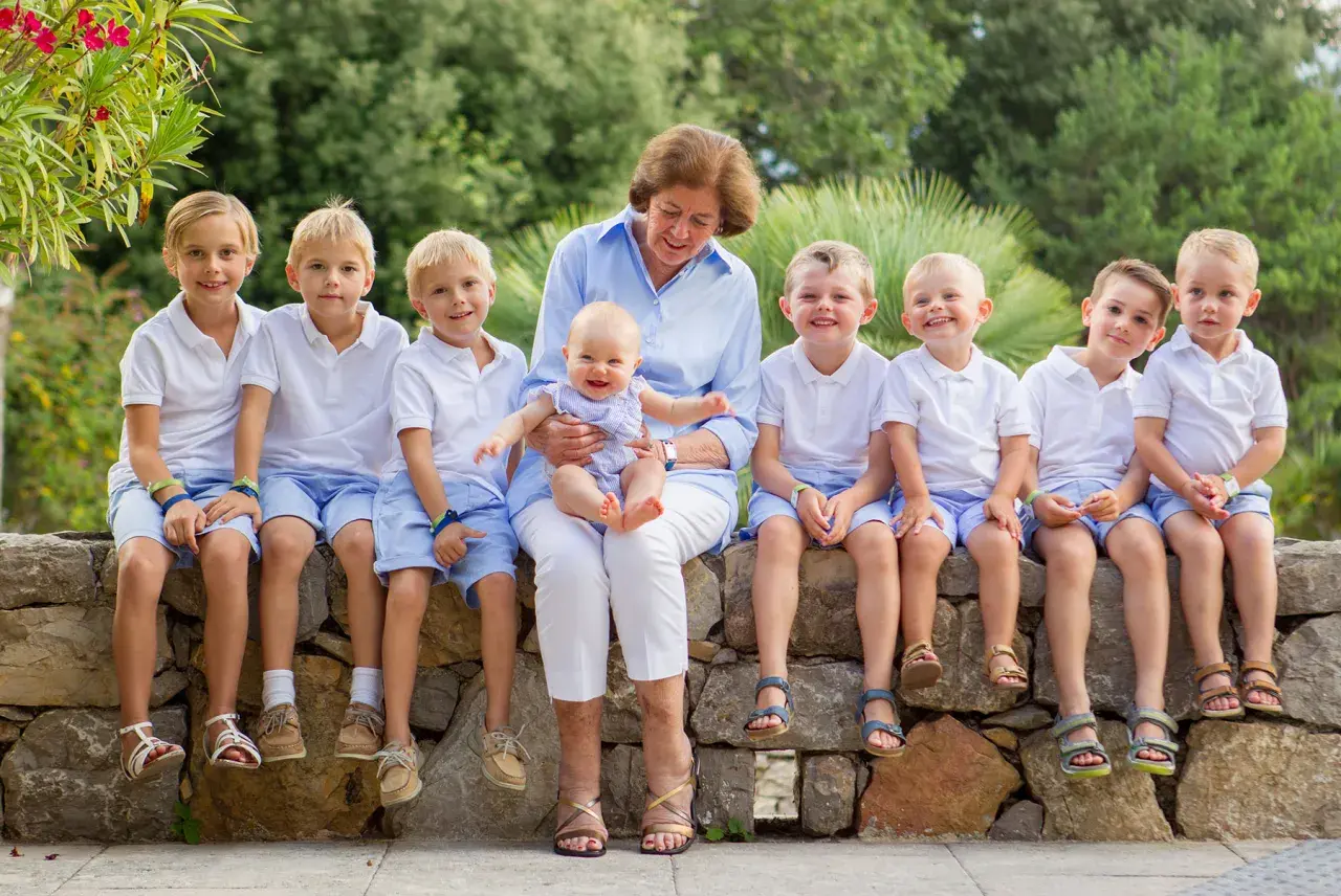 Gammelmormor eller gammelfarmor sitter med ett leende och håller ett litet barn. Runtomkring sitter åtta barn i vita tröjor och blå shorts, alla glada och förväntansfulla.