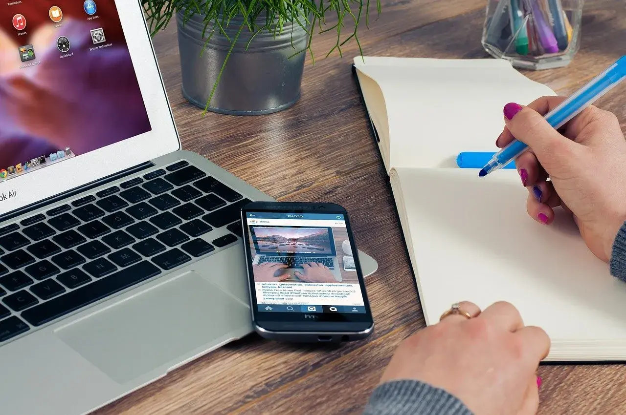 Person skriver i anteckningsbok vid laptop och mobil. Ett bra exempel på en problemformulering kan vara att hitta effektiva metoder för distansarbete.