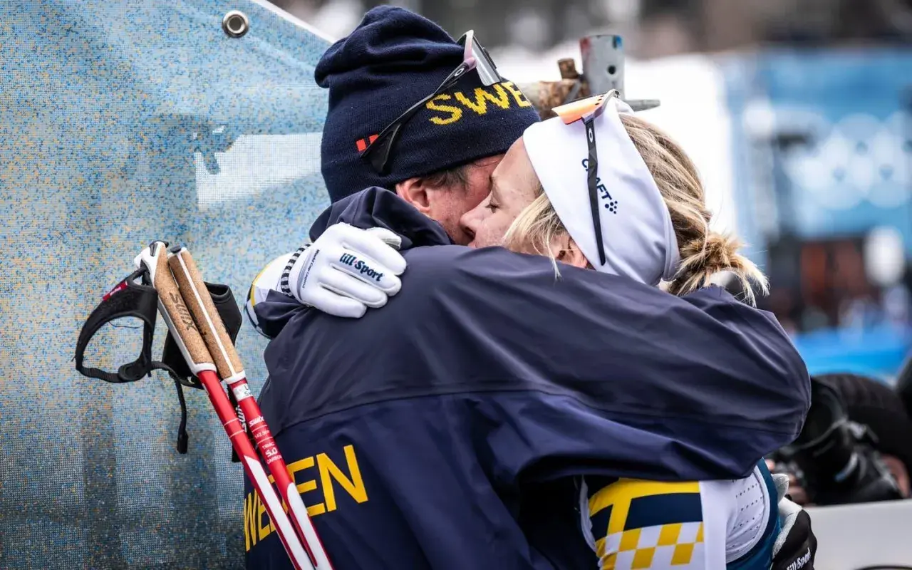 Två skidåkare i svenska färger kramas. Mannen har mössa med texten "SWE" och kvinnan har pannband. De står fram emot varandra i en varm omfamning.