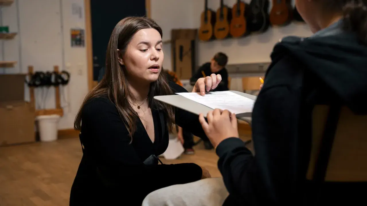 En lärare förklarar hur man skriver en rapport för en elev. I bakgrunden hänger gitarrer.