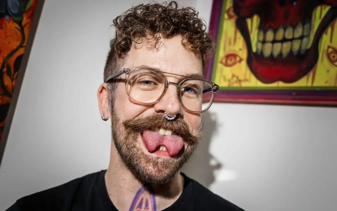 Man med glasögon, mustasch och septumring visar upp sin kluvna tunga. Han verkar tala med kluven tunga, vilket ger ett unikt uttryck.