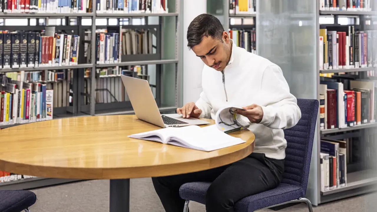 Man sitter vid ett runt bord i ett bibliotek, med en laptop och böcker. Han läser i en bok och funderar på hur citerar man.