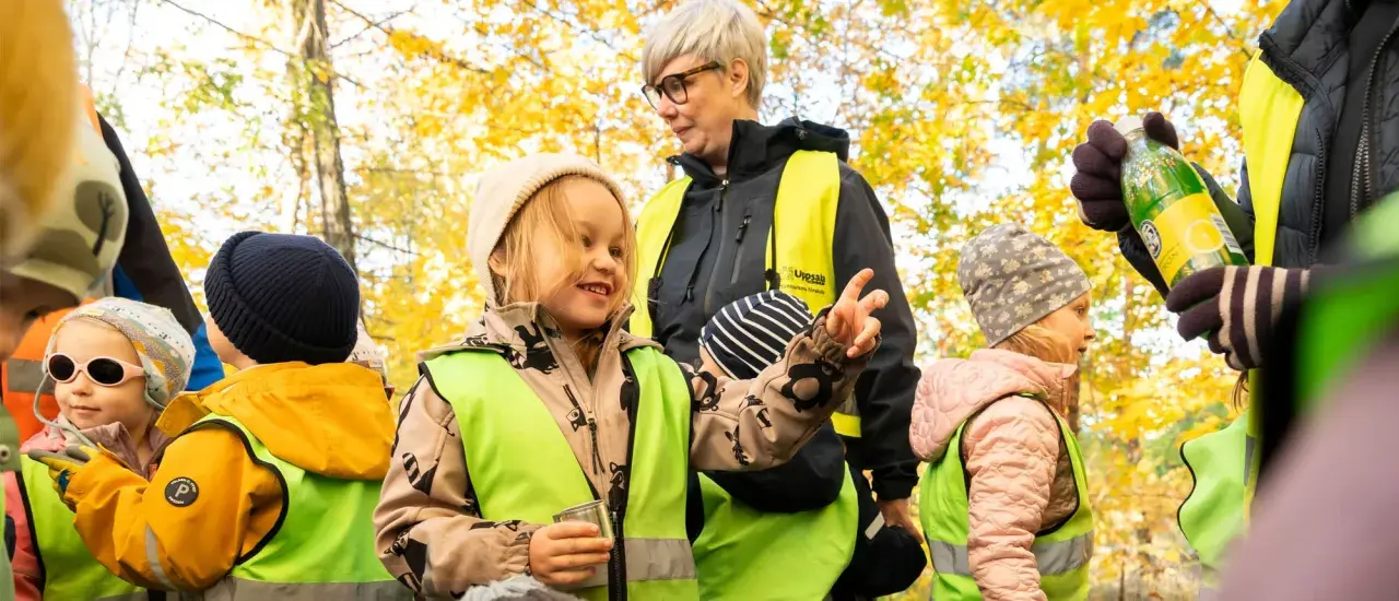 Barn i reflexvästar och mössor i höstskogen. En vuxen deltar i leken med en flaska.