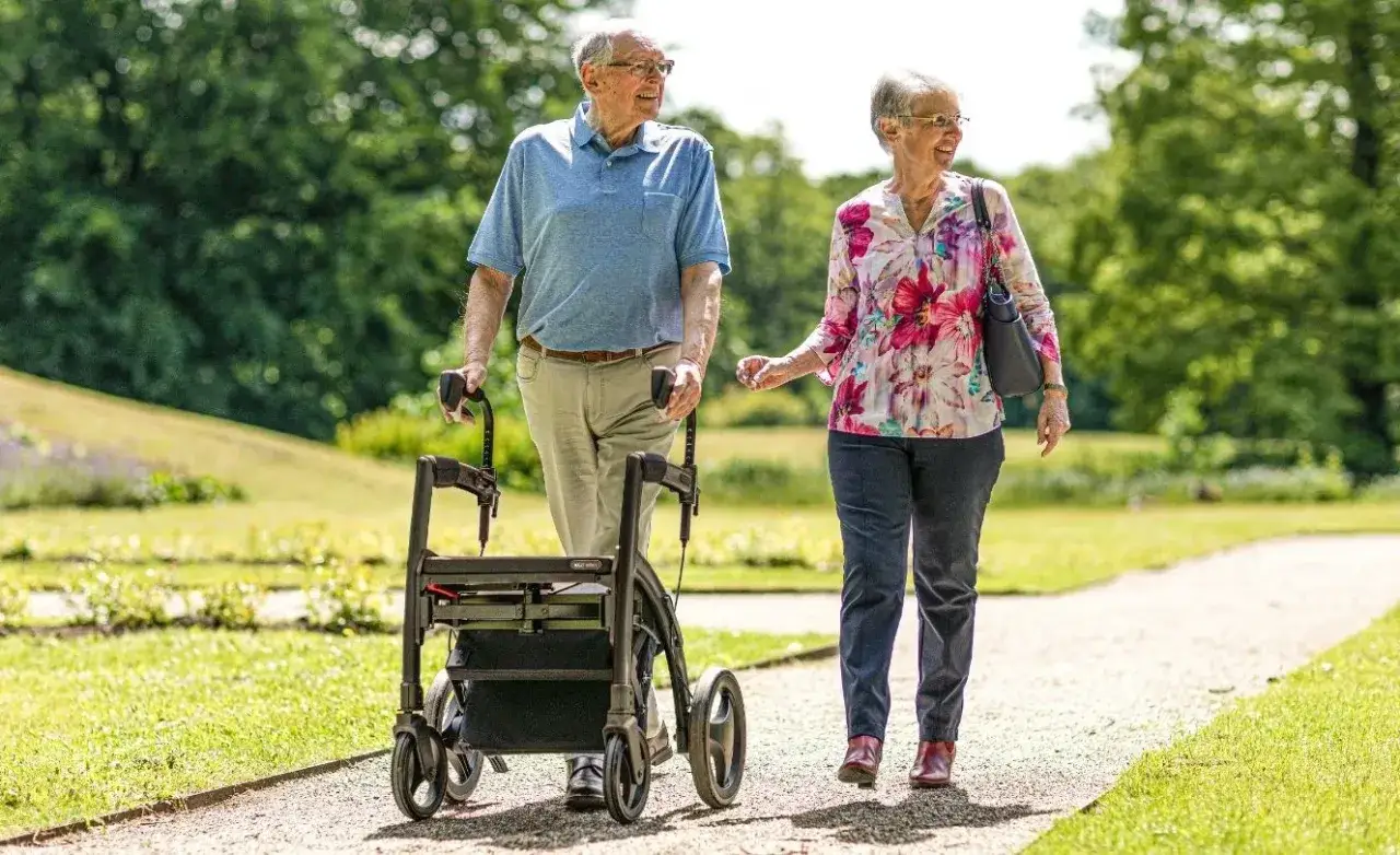 Ett äldre par promenerar på en grusväg i en park. Mannen använder en rollator för stöd.